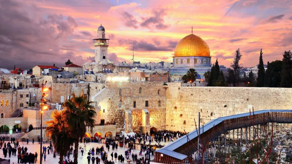 A stunning view of the Western Wall in Jerusalem at sunset, with worshippers gathered in prayer. The golden Dome of the Rock shines in the background, illuminated by the warm hues of pink, orange, and purple in the sky. The historic stone architecture of the Old City contrasts with the modern footbridge leading to the Temple Mount, symbolizing the deep religious and cultural significance of this sacred site. Lush palm trees and glowing streetlights add to the serene and spiritual atmosphere.