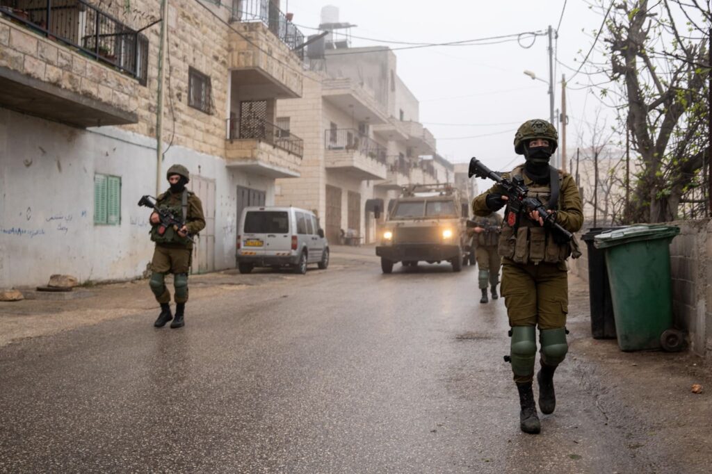 Soldiers patrol street with armored vehicle in background.