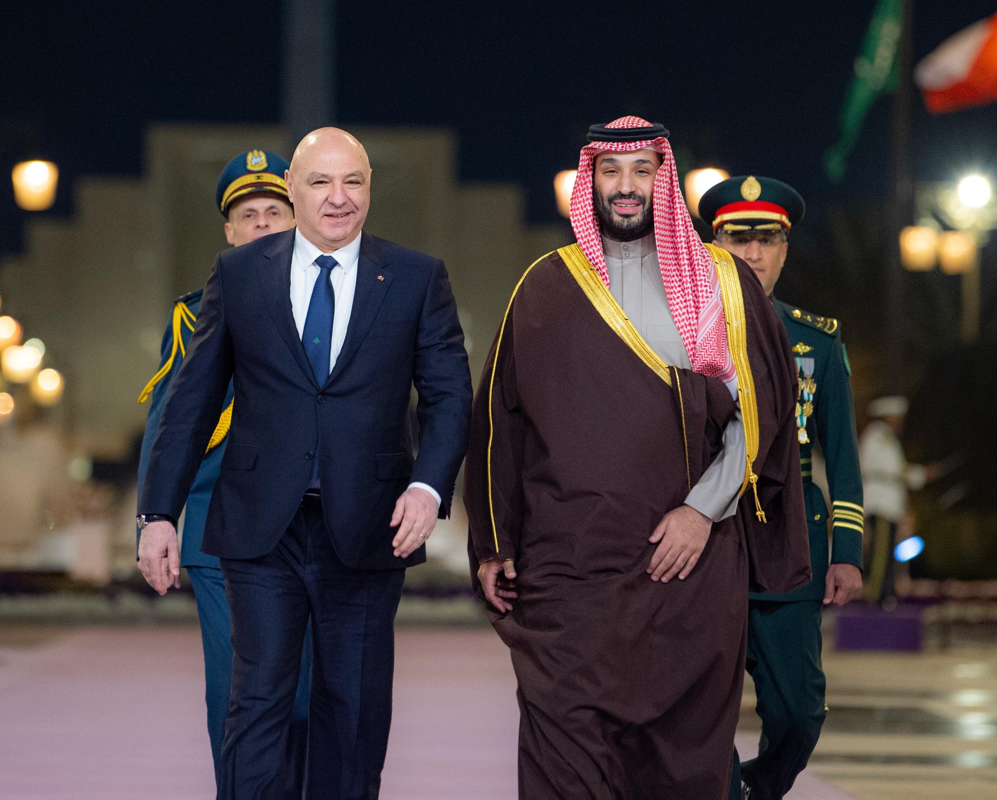 Two men walking in formal attire, uniformed guards behind.